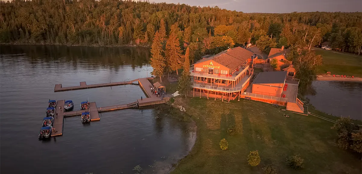 Eine am See gelegene Lodge, umgeben von Herbstbäumen, mit Stegen, die in das ruhige Wasser hineinragen, und mehreren Booten, die am Ufer vertäut sind.