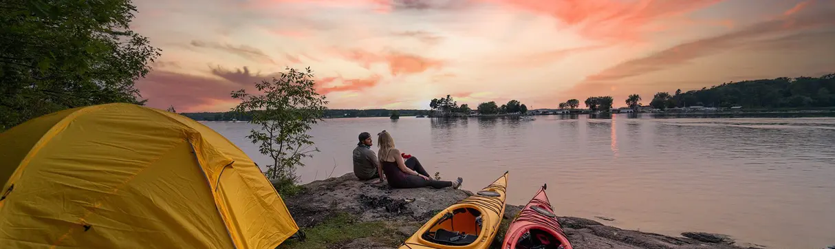 A couple sits on a rock in front of their tent and kayaks to watch the sunset over the St. Lawrence River.