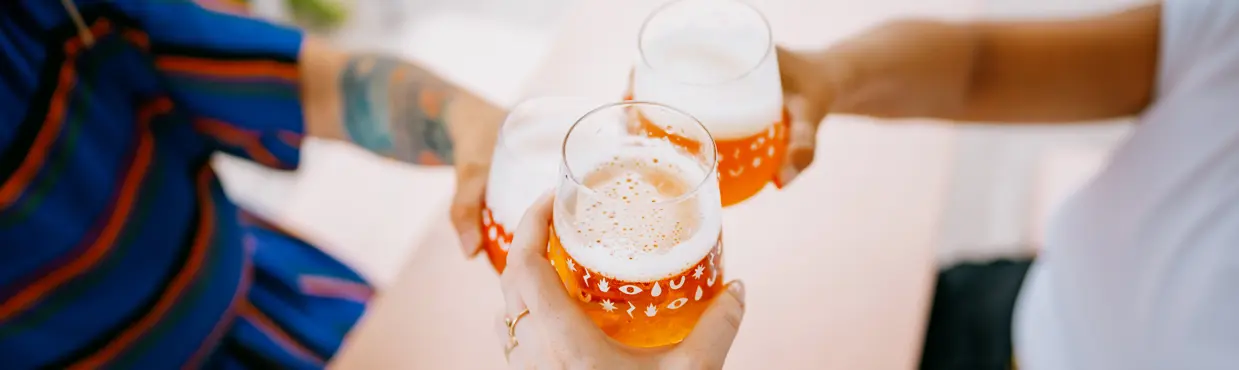 Three people clinking glasses of beer at a pink table, with playful patterns on the glasses in a relaxed outdoor setting.