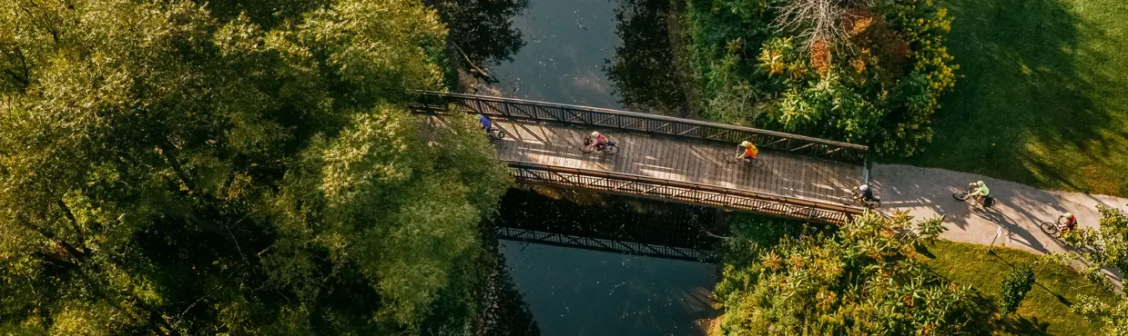  深い緑と広々とした芝生に囲まれた静かな川にかかる木製の橋を渡るサイクリストたちの空中写真。