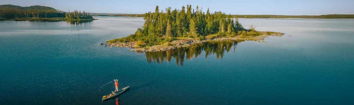 Une personne qui fait de la planche à pagaie sur un lac paisible, qui lance une ligne de pêche à l’eau près d’une petite île couverte d’arbres, sous un ciel bleu clair.