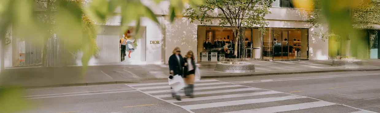 쇼핑백을 들고 횡단보도를 건너는 세 사람. 흐릿한 녹색 잎사귀에 둘러싸인 가운데, 고급 매장 앞.