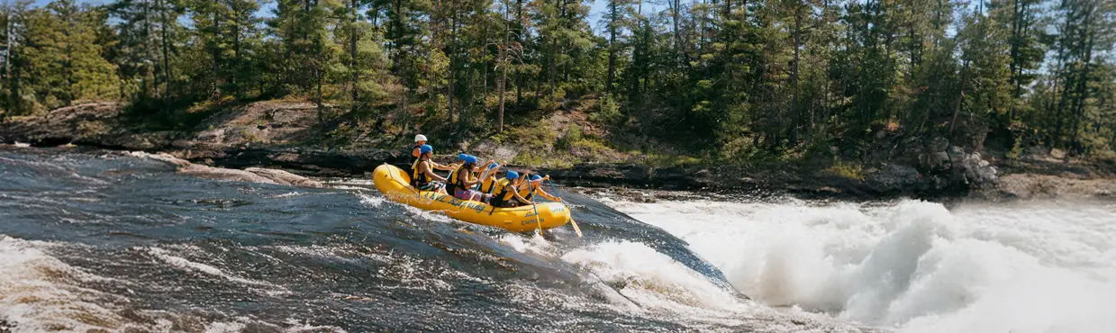 A group of rafters wearing life jackets and helmets paddling a yellow inflatable raft down the Ottawa River, surrounded by a dense forest.