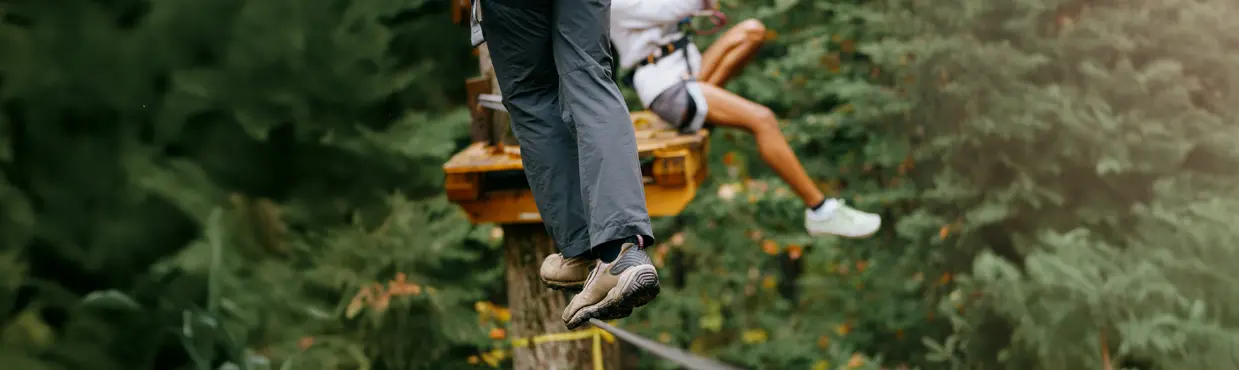 A person slacklining in safety gear while another, harnessed, prepares to zipline from a wooden platform in Algonquin Provincial Park.