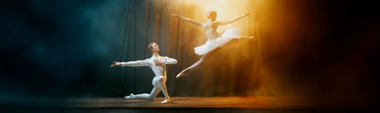  Eine Ballerina springt in einem weißen Kostüm über eine schwach beleuchtete Bühne, während ein männlicher Tänzer in einer dramatischen Ballettvorstellung kniet und die Arme ausstreckt.