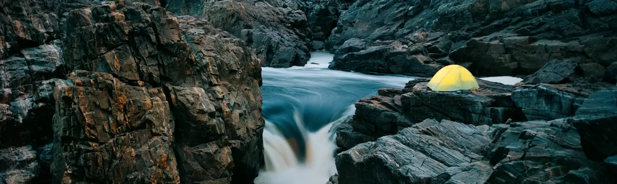 A glowing tent set up on rocky cliffs above Missinaibi River rapids near Sault Ste. Marie in Algoma, surrounded by forest.