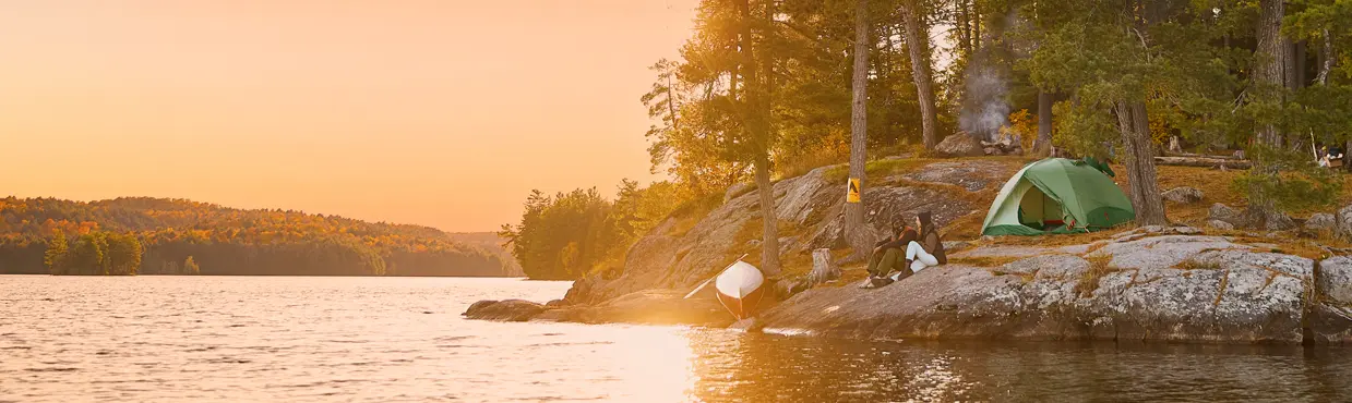 Ein Campingplatz am Opeongo Lake mit Zelt und Kanu auf einem Felsvorsprung, umrahmt von hohen Kiefern und goldenem Sonnenuntergangslicht.
