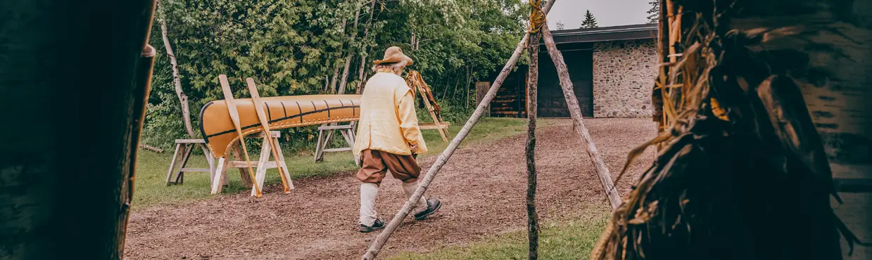 Une personne en costume d'époque passant devant un canot exposé, vu de l'intérieur d'une structure autochtone avec un foyer sur l'herbe.