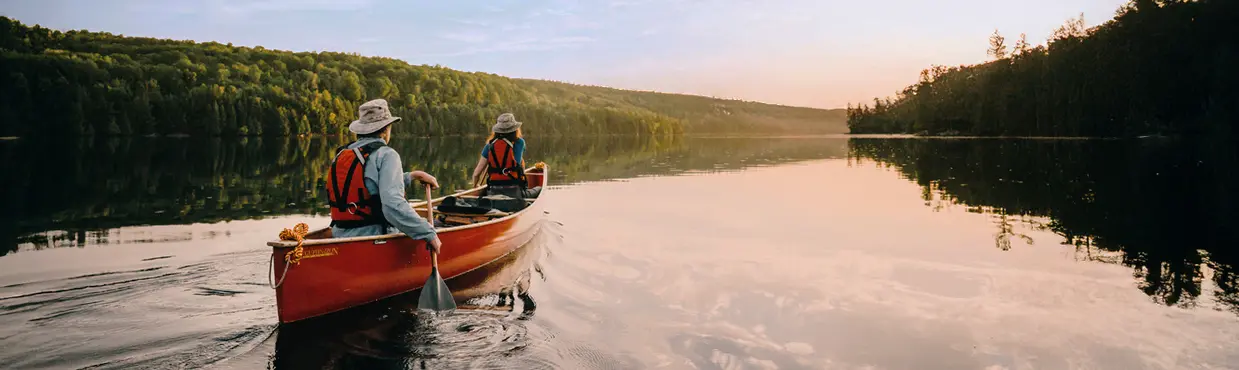 Dos personas remando en una canoa roja por las tranquilas aguas dulces de Parry Sound, rodeadas de un exuberante bosque bajo un cielo suave y nublado.