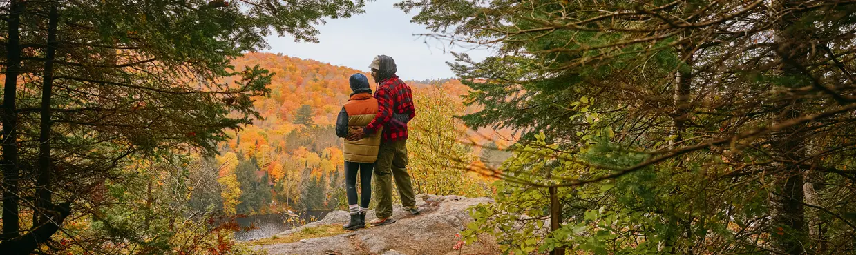 Two people in an embrace on a rocky ledge, overlooking Oxtongue Lake and a valley of vibrant fall foliage in Ontario.