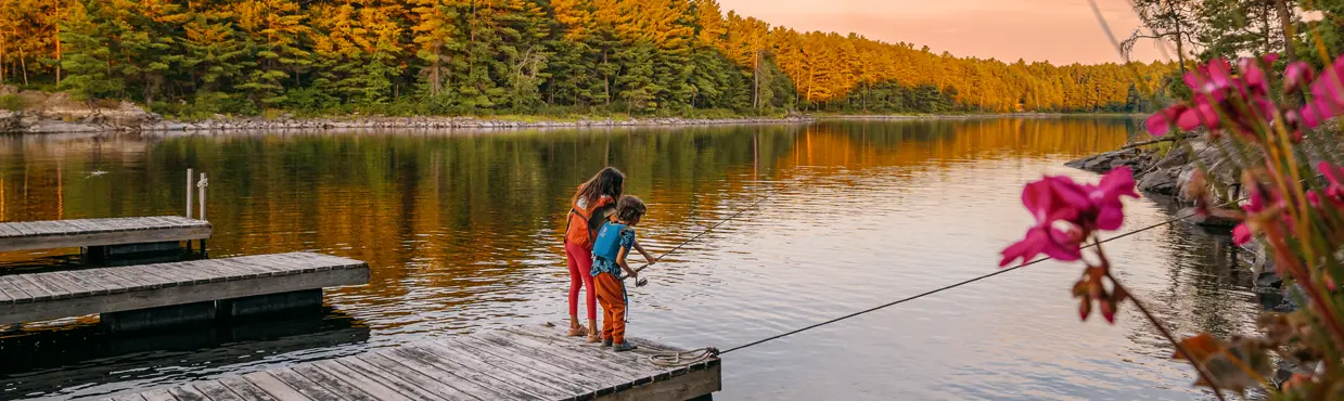 프렌치 강에 있는 크레인 로차벤 롯지 근처의 일몰 무렵, 나무 부두에서 낚시를 하는 아이들. 숲과 고요한 물에 둘러싸여 있습니다.