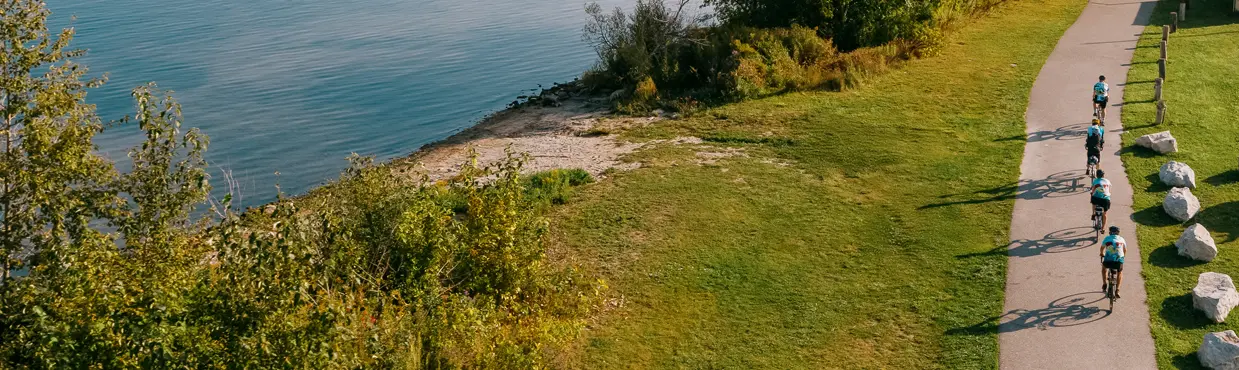 オンタリオ州コリングウッドのジョージアン湾沿いのウォーターフロントの小道をサイクリストたちが走り、遠くに静かなマリーナと帆船の眺めを楽しみます。