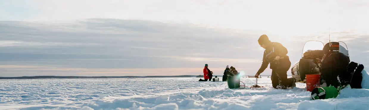 Personne pêchant sur la glace sur le lac Nipissing au lever du soleil, agenouillée à côté d'un équipement et d'une motoneige avec d'autres pêcheurs en arrière-plan.