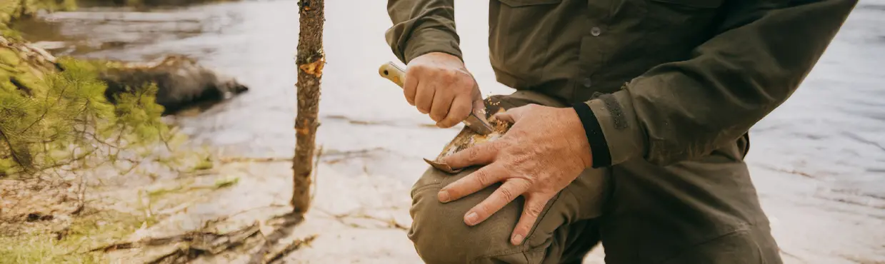 와바키미 주립공원의 호수 옆에서 장작을 썰기 위해 칼을 사용하는 사람의 클로즈업. 근처에 작은 나무줄기와 가지가 있다.
