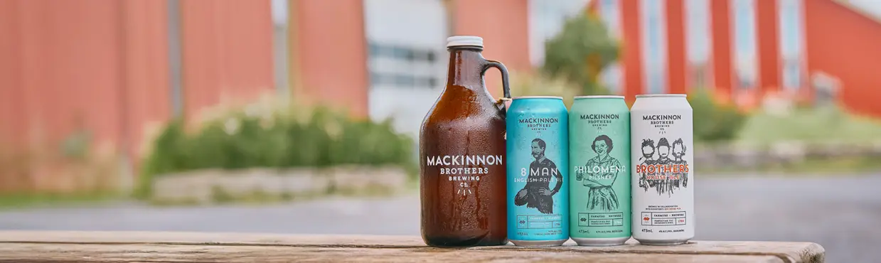 Une bouteille de bière et trois canettes de Mackinnon Brothers (8 Man, Philomena et Brothers House Ale) sur une table, granges en arrière-plan.