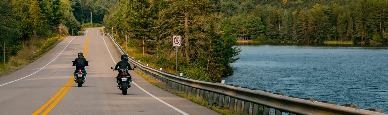 Zwei Motorradfahrer fahren an einem sonnigen Tag auf einer ruhigen Uferstraße durch die Waldlandschaft der Ontario Highlands.