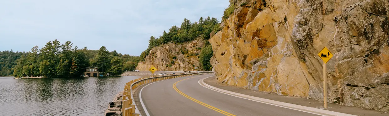 A winding road along a rocky cliffside beside a calm lake, with a yellow road sign warning of falling rocks, in Ontario's District of Parry Sound.
