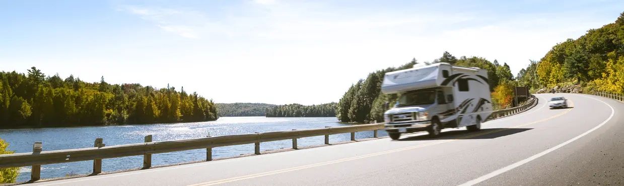  알곤퀸 공원의 숲으로 둘러싸인 햇살 가득한 호숫가 길을 따라 RV를 타고 달리는 모습.