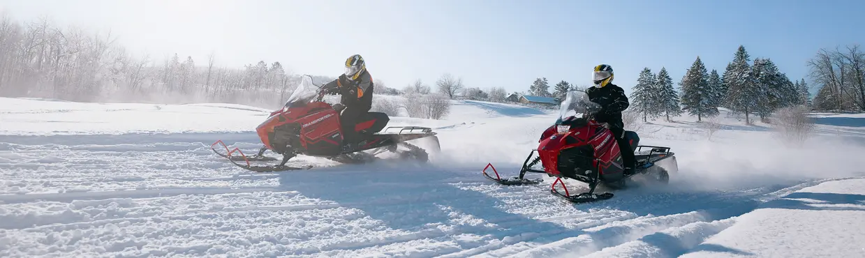 Two people riding red snowmobiles through powdery snow on a bright winter day at Deerhurst Resort.