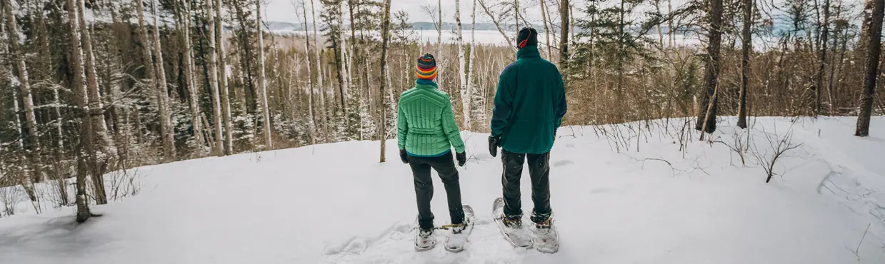 サドベリー近郊のウィンディ レイク州立公園の雪道をスノーシューで歩く 2 人が、高い木々の間から凍った湖を眺めている。