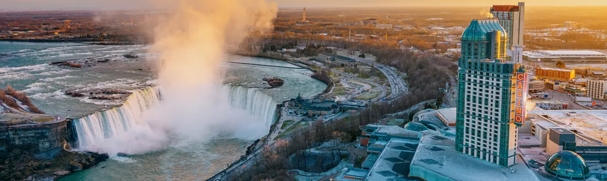 Aerial view of Horseshoe Falls and downtown Niagara Falls at sunset, with mist rising and rooftops dusted in snow.