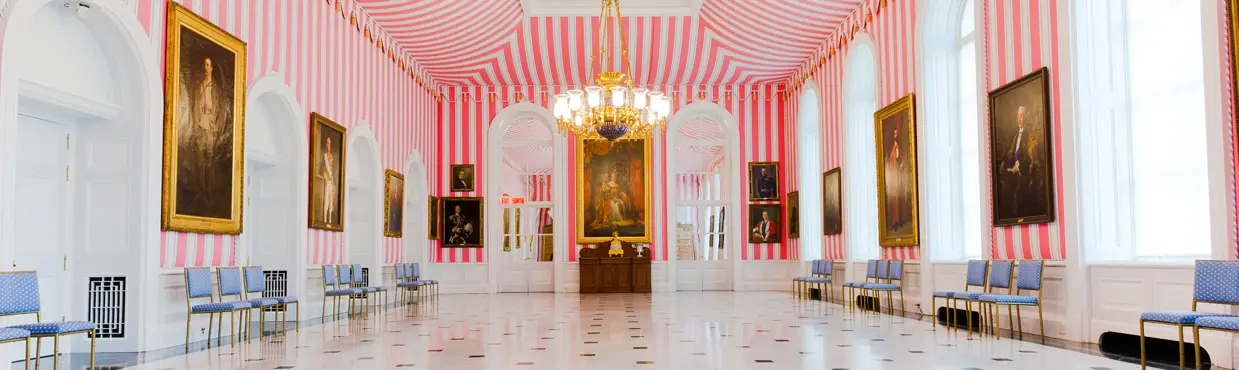  Innenansicht des Zeltraums im Rideau Hall in Ottawa mit kräftig rosa-weiß gestreiften Wänden, goldenen Kronleuchtern und gerahmten Porträts, die den Raum säumen.