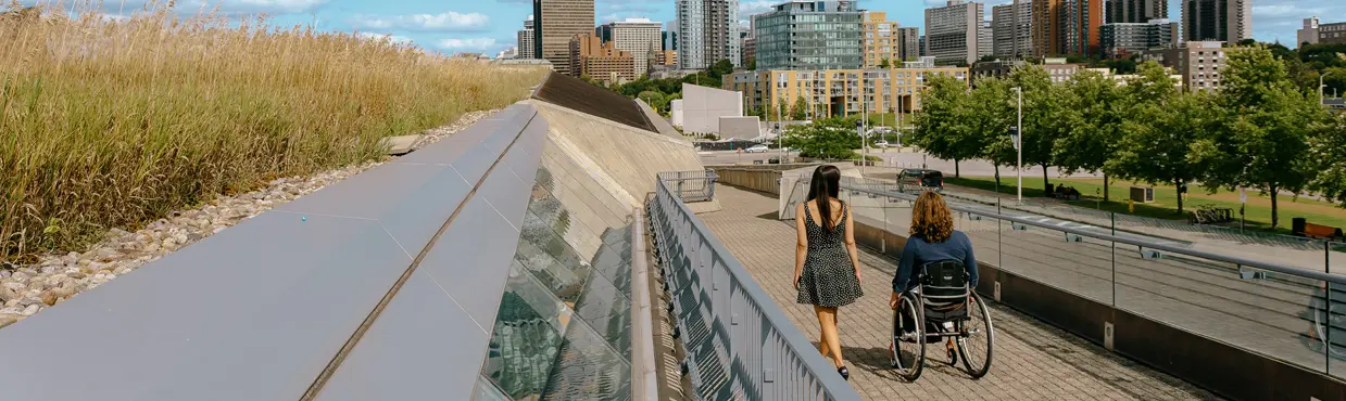 밝은 날, 경사진 녹색 지붕이 있는 도시 산책로를 따라 휠체어를 탄 두 사람이 걷고 있습니다. 배경에는 오타와 도시의 스카이라인이 보입니다.