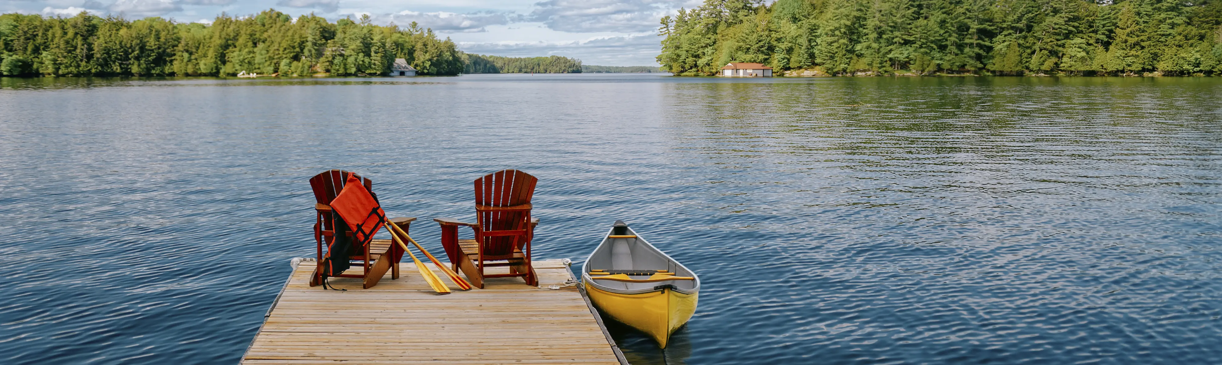 Two Adirondack chairs on a wooden dock overlooking the blue waters of a Muskoka lake in Ontario, Canada. 