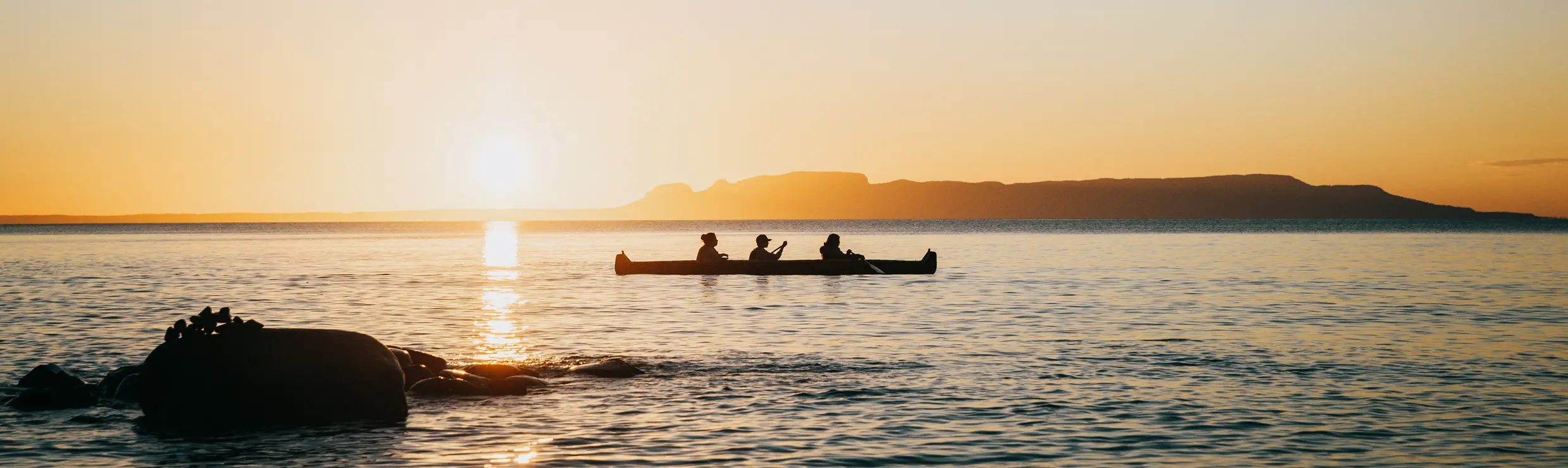 Three people paddling a long canoe in an amber sunset on a still blue lake.