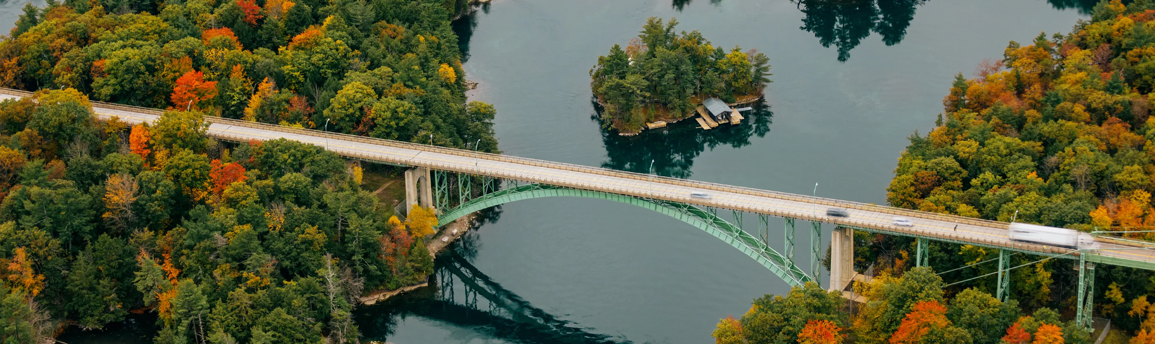 Toma aérea de un puente largo que se extiende sobre un lago tranquilo y áreas con árboles frondosos que se vuelven anaranjados.