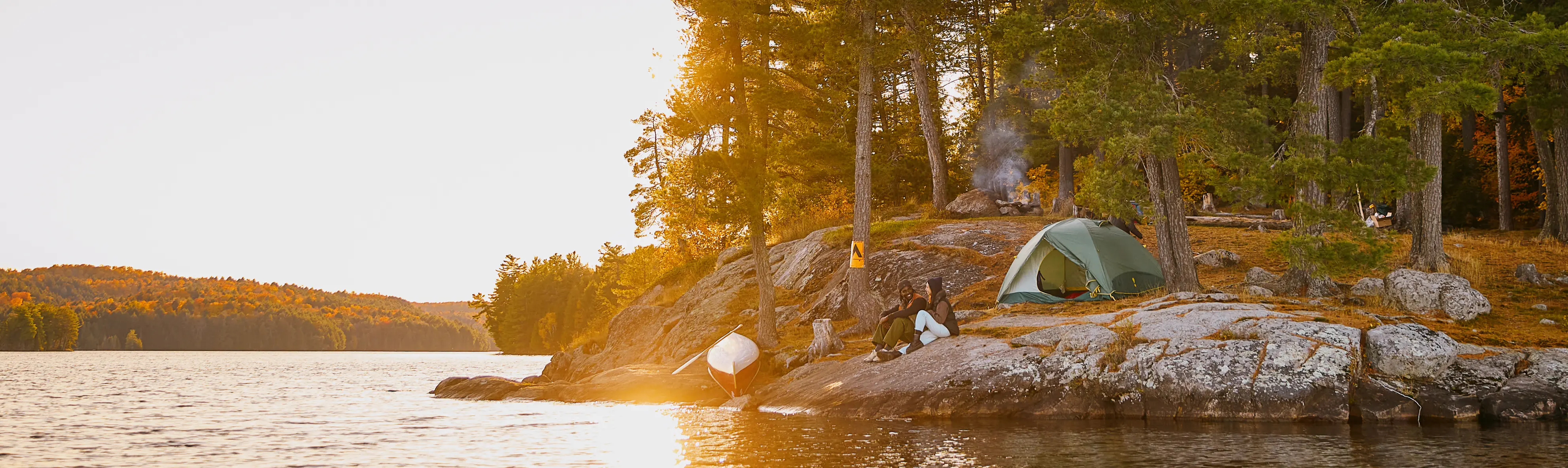Ein Campingplatz auf einer Insel, umgeben von hohen Bäumen, bei Sonnenaufgang, während ein Camper vor seinem grünen Zelt sitzt.
