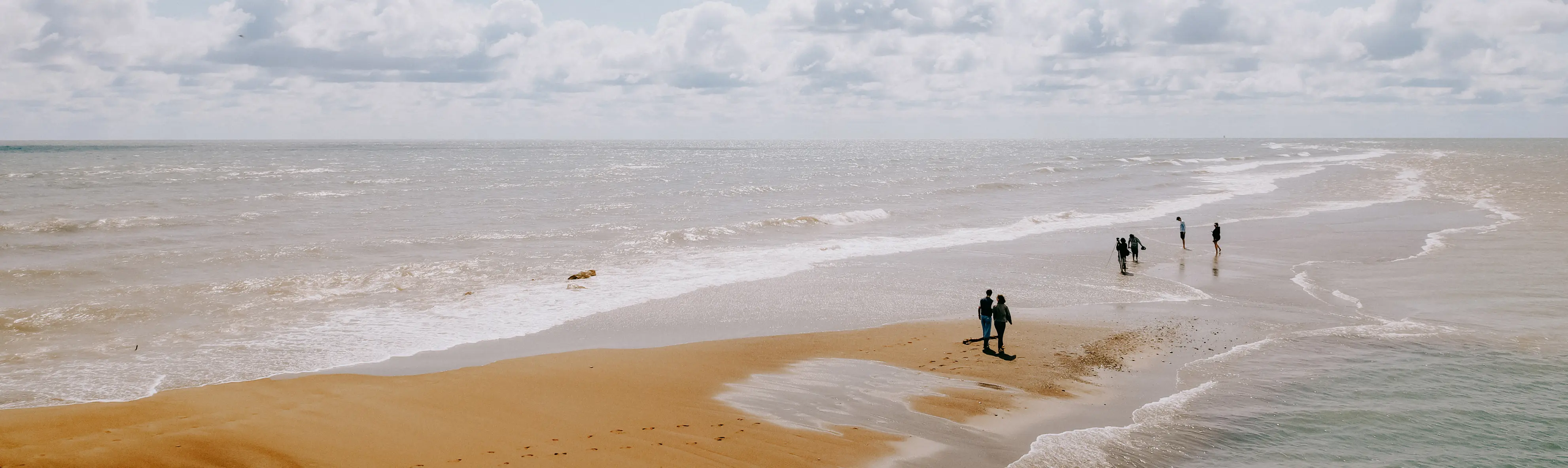 People strolling along a red sandbar stretching into the lake, with gentle waves and clouds on a bright day.