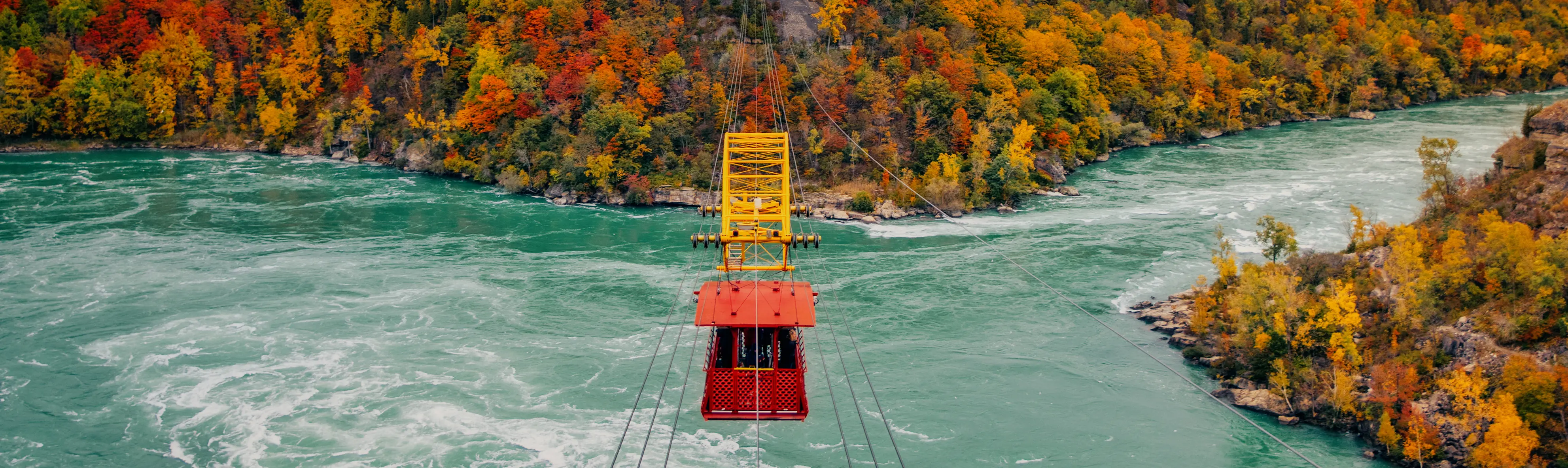 Niagara's red whirpool aero car riding across the rushing falls, heading towards colourful autumn trees.