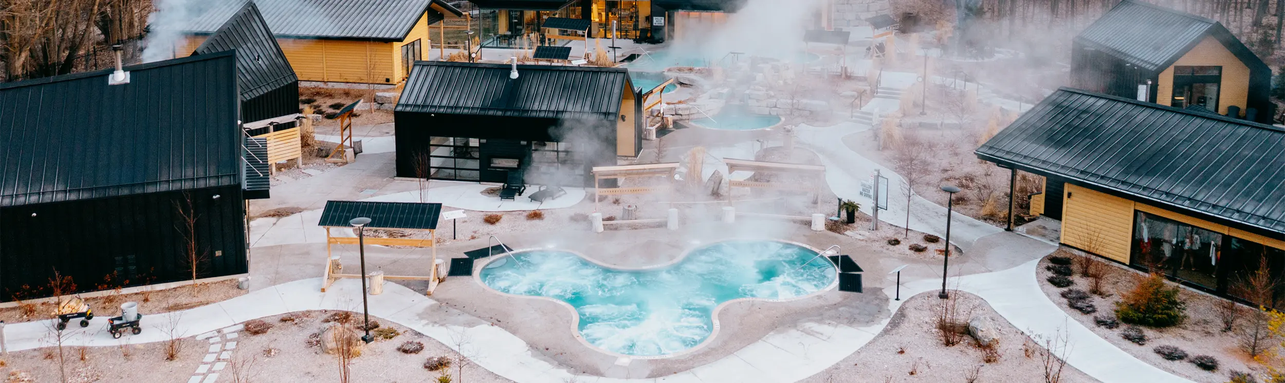 Vapeur s’élevant au-dessus des bassins chauds et froids, des saunas et des hammams d’un spa nordique extérieur entouré d’arbres à la fin de l’automne.