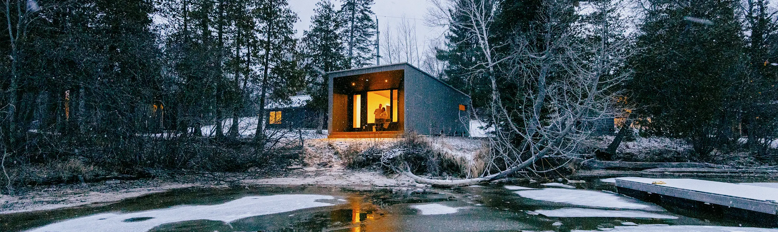 A modern cabin with glowing windows overlooks a frozen lake, surrounded by snowy ground and bare trees at twilight.