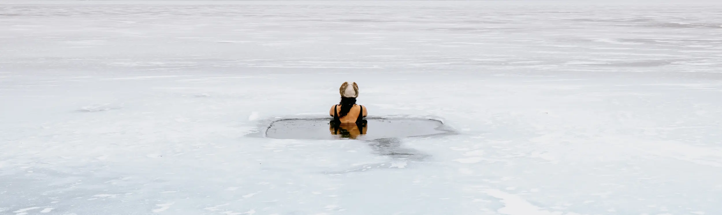Eine Frau nimmt ein kaltes Bad in einem Loch, das in einen zugefrorenen See gegraben wurde, umgeben von schneebedeckten Hügeln unter einem bedeckten Himmel.