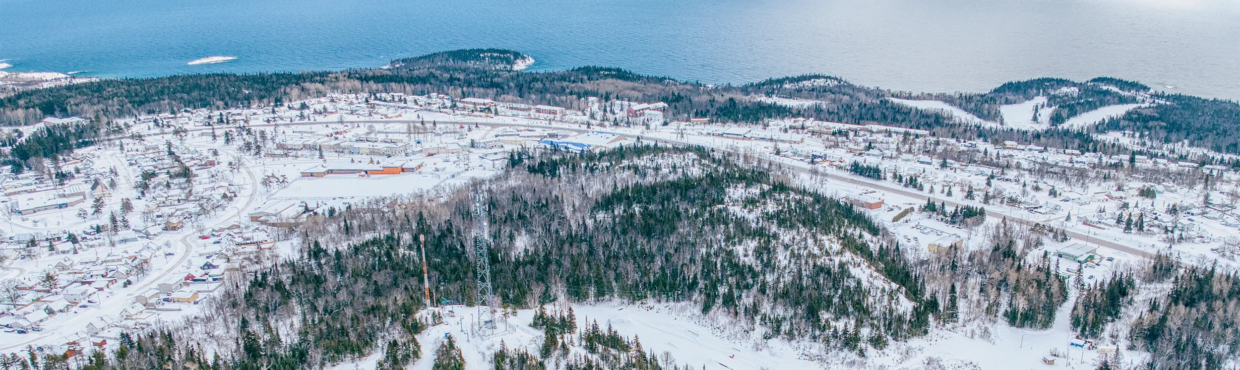 大きな水域に沿った小さな町を見下ろす、雪に覆われたスキー場と森林に覆われた尾根、曇り空の下の曲がりくねったスキーコースと遠くの海岸線を空から眺めた冬の眺め。