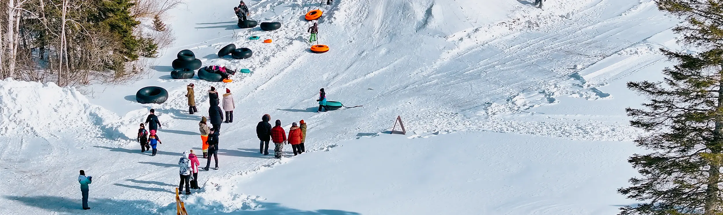 Gente haciendo tubing en una colina nevada en una zona boscosa, algunos caminando y otros jugando en la nieve.