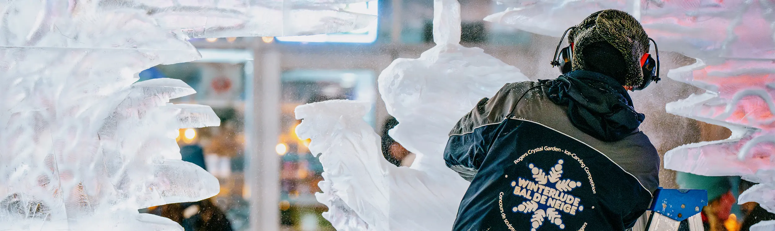 夜間に屋外で大きな氷の彫刻を彫る芸術家。防護服を着用しており、背景では観客が見守っている。