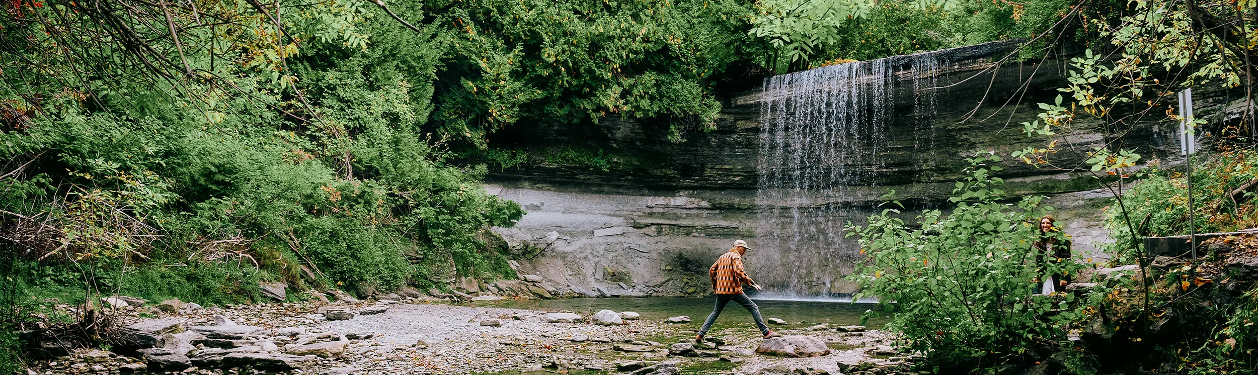 A person walking across rocks in a creek in front of a small waterfall surrounded by lush green trees.