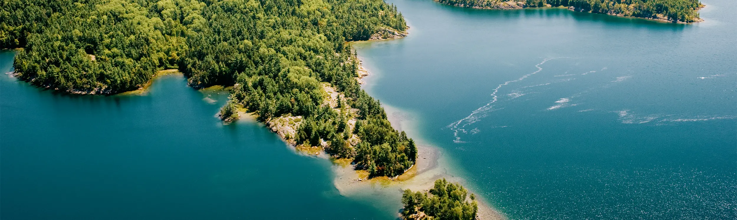 Luftaufnahme von bewaldeten Inseln, umgeben von ruhigem, tiefblauem Wasser unter klarem Himmel.
