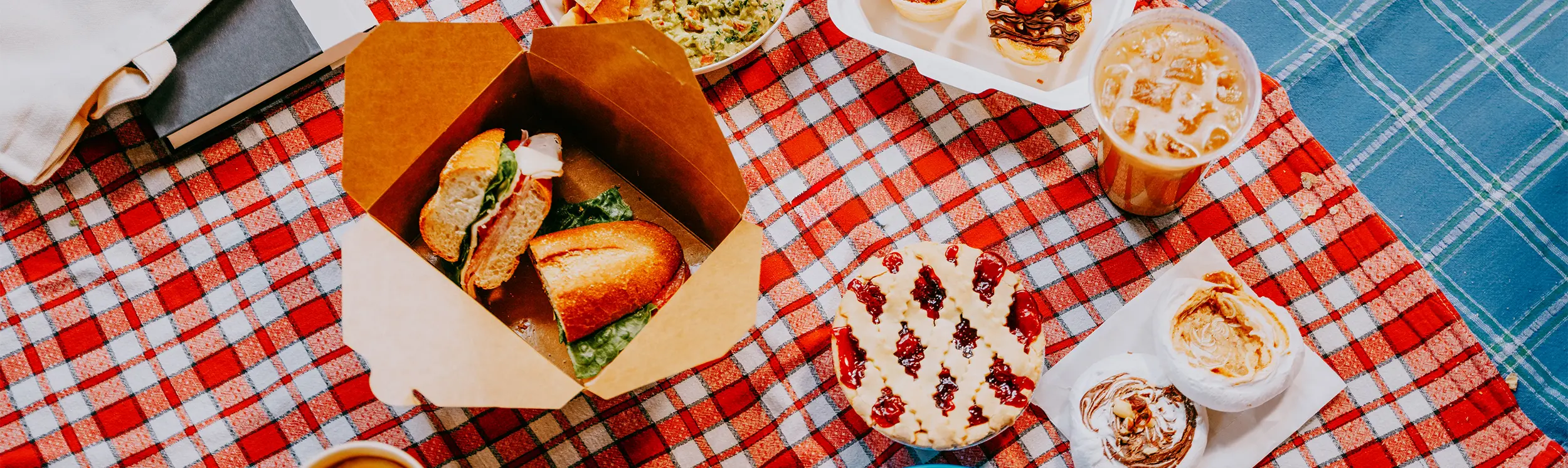 체크무늬 담요 위에 차려진 피크닉 음식들. 샌드위치, 과카몰리를 곁들인 감자칩, 디저트, 음료 등이 준비되어 있다.