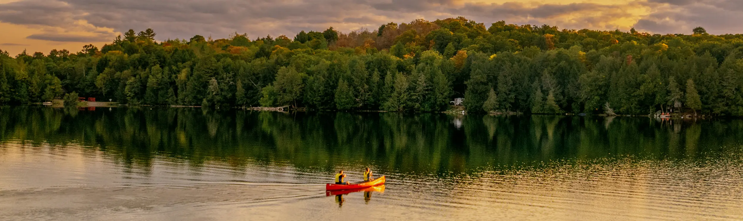   Two people paddling a red canoe across a calm lake at sunset with forest along the shoreline.
