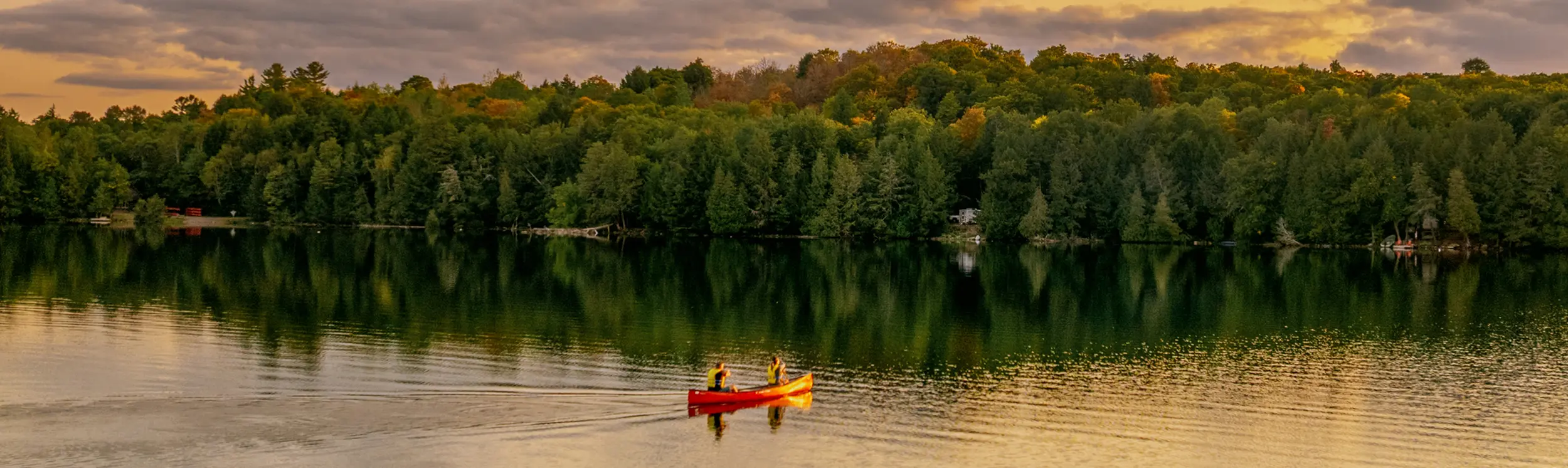 Deux personnes pagayent dans un canoë rouge sur un lac calme au coucher du soleil, avec une forêt le long de la rive.