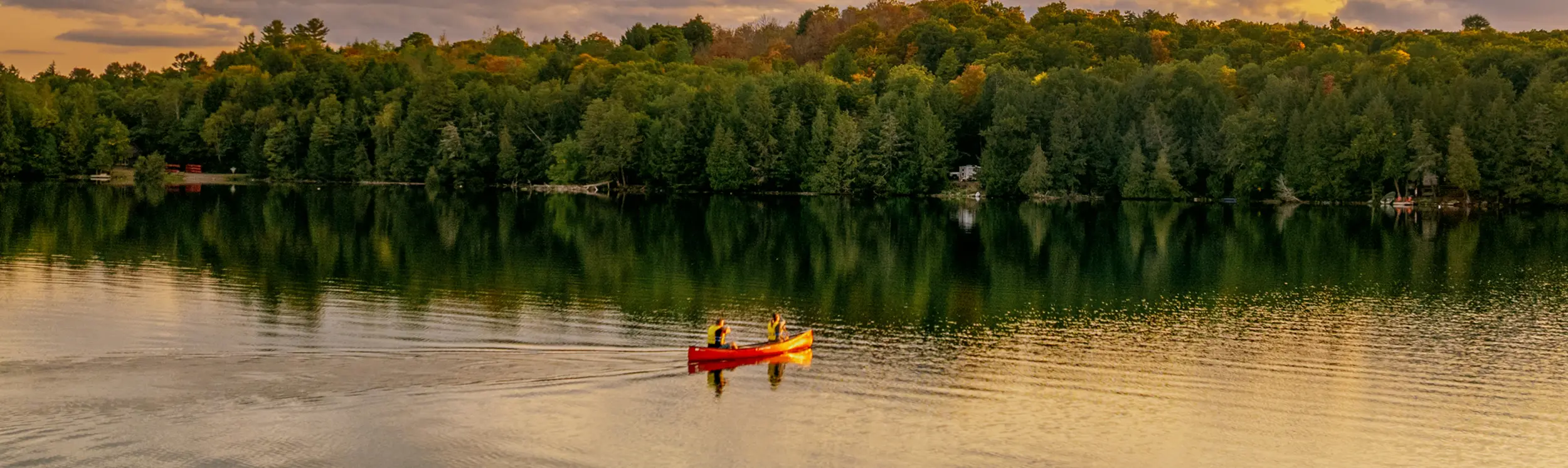 Two people paddling a red canoe across a calm lake at sunset with forest along the shoreline.