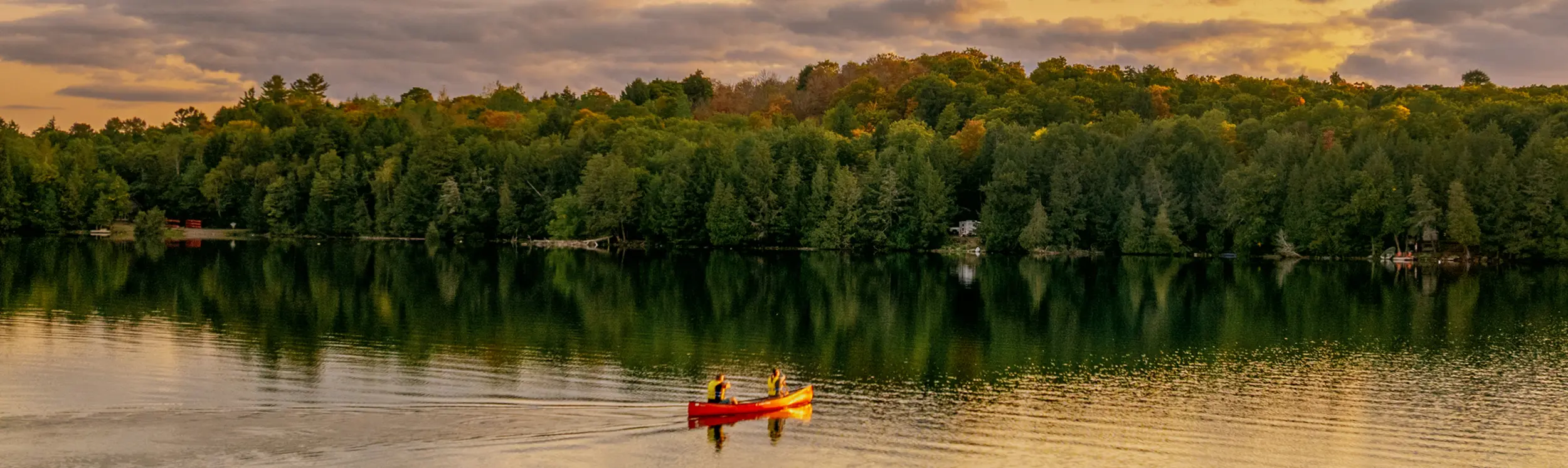 Dos personas remando en una canoa roja a través de un lago tranquilo al atardecer, con un bosque a lo largo de la orilla.