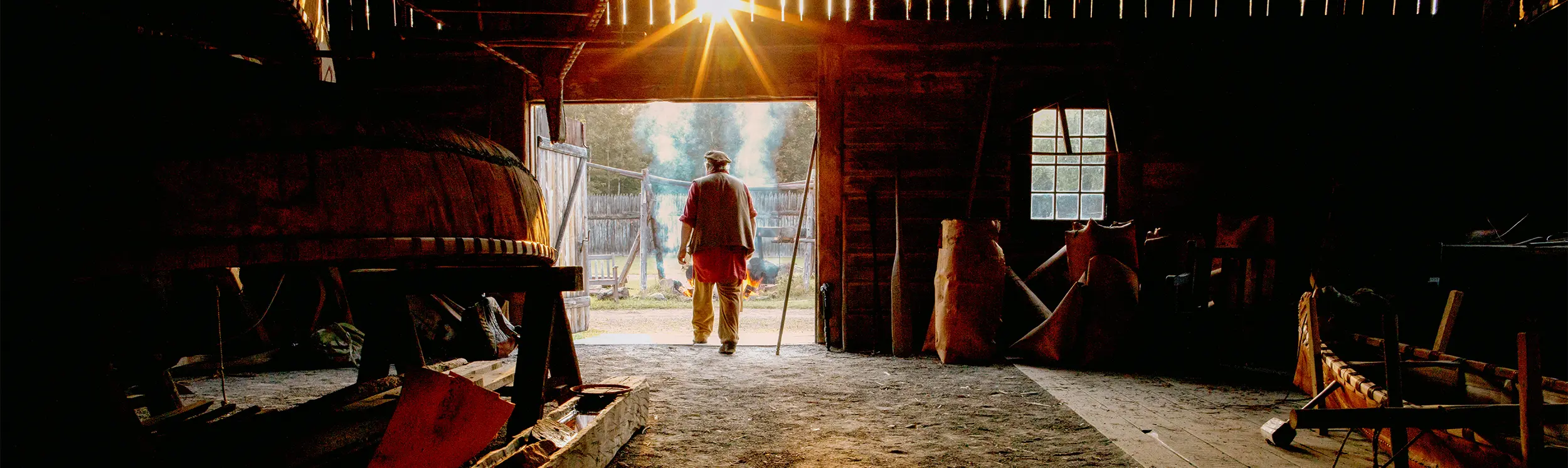 A silhouetted figure walks out of a sunlit barn doorway, surrounded by canoes and woodworking tools.