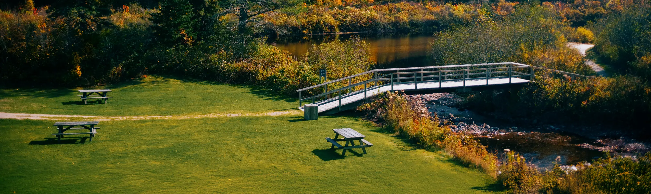 Eine Fußgängerbrücke über einen Bach in einem Tal mit Herbstbäumen und Picknicktischen.