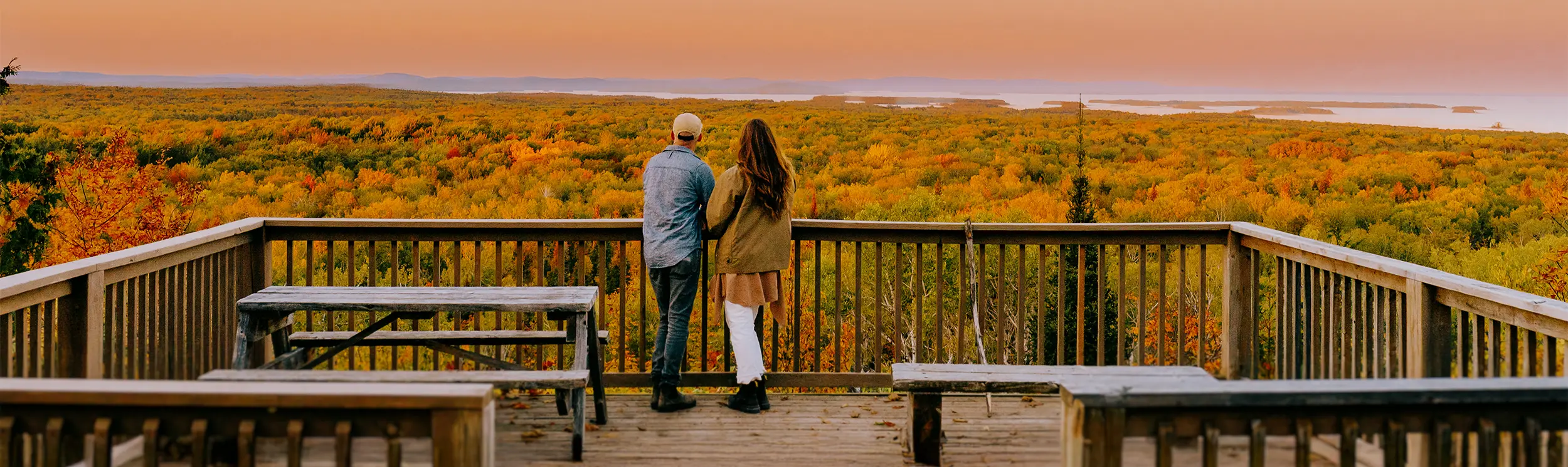 K2  Two people stand on a lookout deck with benches overlooking an autumn forest and distant water at sunset.