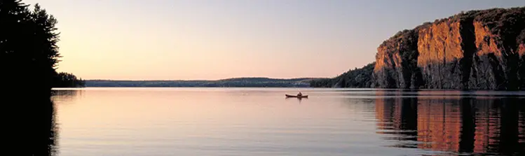 Cliffs overlook a serene lake at dusk with one lone kayaker.